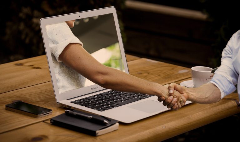 Der Arm einer Person kommt aus dem Bildschirm eines Laptops heraus und schüttelt die Hand einer anderen Person, die vor ihrem Laptop an einem Tisch sitzt. Online Verbundenheit kann die Offline Verbundenheit stärken.