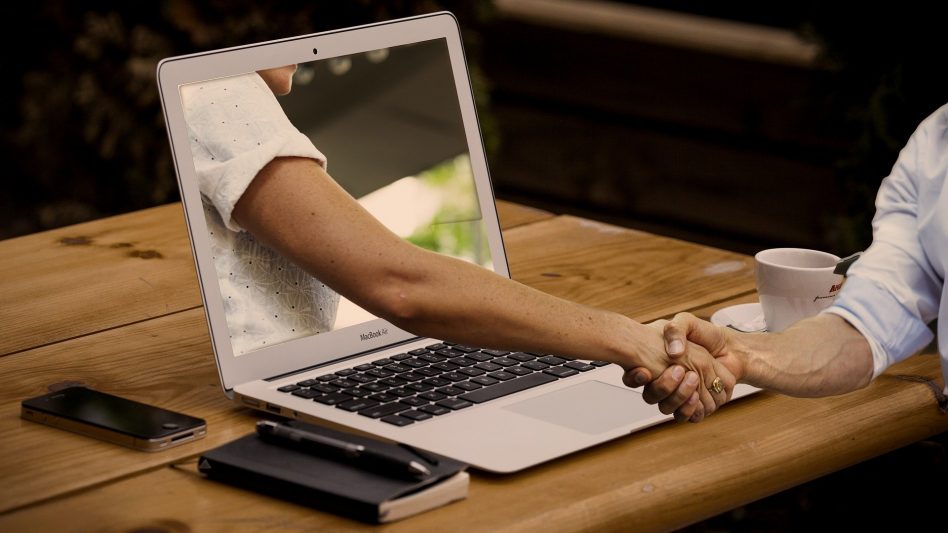 Der Arm einer Person kommt aus dem Bildschirm eines Laptops heraus und schüttelt die Hand einer anderen Person, die vor ihrem Laptop an einem Tisch sitzt. Online Verbundenheit kann die Offline Verbundenheit stärken.